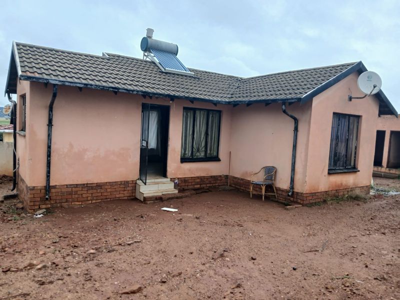 Rear view of a single-story house featuring a tiled roof, solar water heater, and brick plinth foundation, set on an unpaved yard.