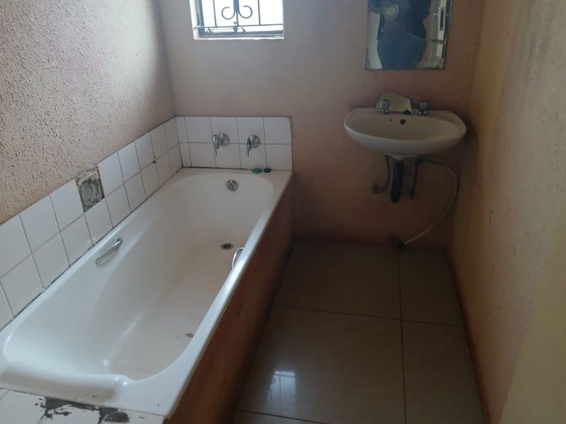 A dated bathroom featuring a built-in bathtub, wall-mounted basin, and tiled finishes, currently requiring cosmetic updates and repairs to fixtures and wall surfaces.