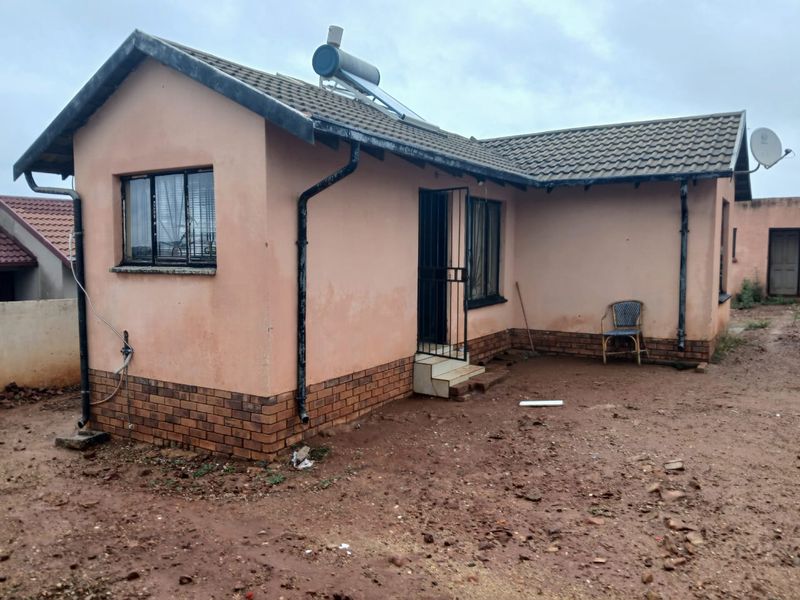 Rear view of a single-story house featuring a tiled roof, solar water heater, and brick plinth foundation with unpaved surrounding grounds.