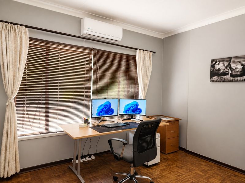 A well-maintained home office featuring parquet flooring, a wall-mounted split-system air conditioner, and large windows with venetian blinds.