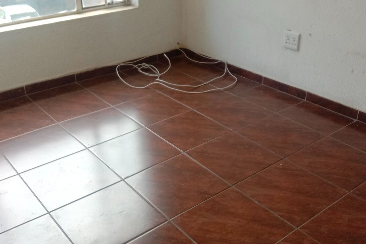 An empty bedroom featuring dark brown tiled flooring, white walls, and a large window, presenting a blank canvas for renovation.