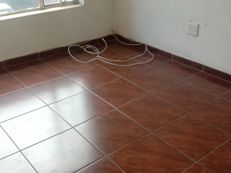 An empty bedroom featuring dark brown tiled flooring, white walls, and a large window, presenting a blank canvas for renovation.