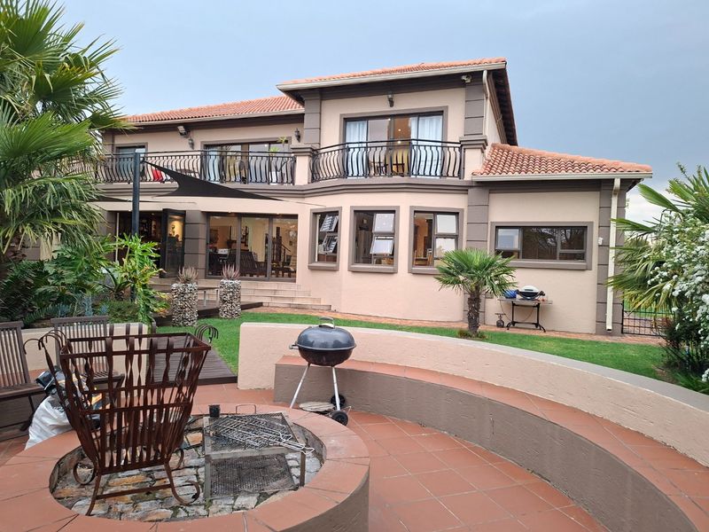 Rear view of a two-story house featuring a spacious terracotta patio, built-in fire pit, curved seating wall, and an upper-level balcony with wrought iron railings.