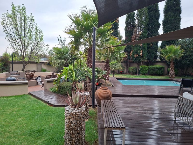 Outdoor entertainment area featuring a swimming pool, extensive timber decking, built-in braai, and shade sails, set within a landscaped garden.