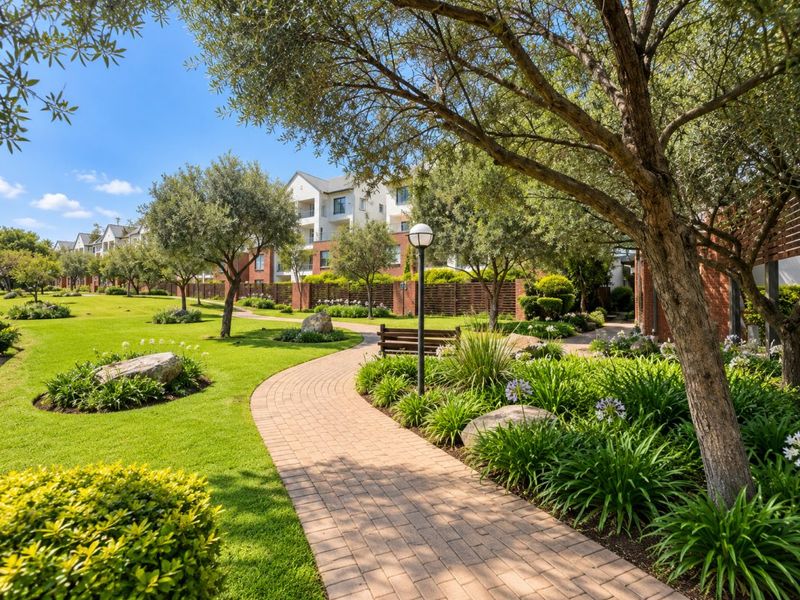 A beautifully landscaped communal garden featuring a paved walkway, mature trees, and well-maintained lawns within a residential apartment complex.