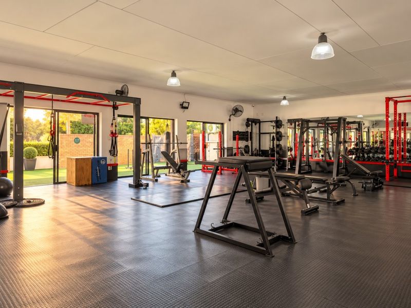 A spacious, well-equipped gym featuring durable rubberized flooring, large sliding glass doors for natural light, and a full-length mirrored wall.