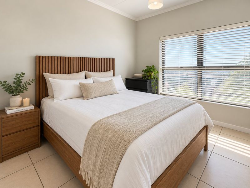 A well-maintained bedroom featuring neutral tiled flooring, large window with venetian blinds, and clean finishes.