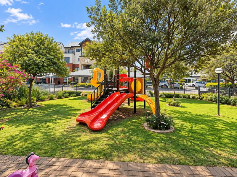 A well-maintained communal garden area featuring a children's playground, manicured lawn, and paved pathways within an apartment complex.