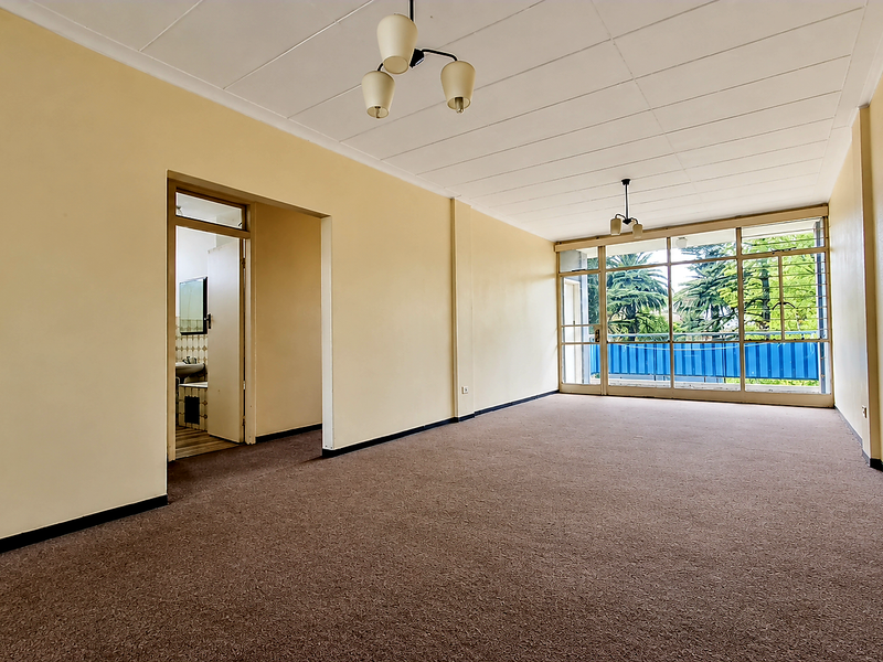 A spacious, empty living room featuring carpeted flooring, dated ceiling tiles, and large glass sliding doors that provide natural light and access to the balcony.