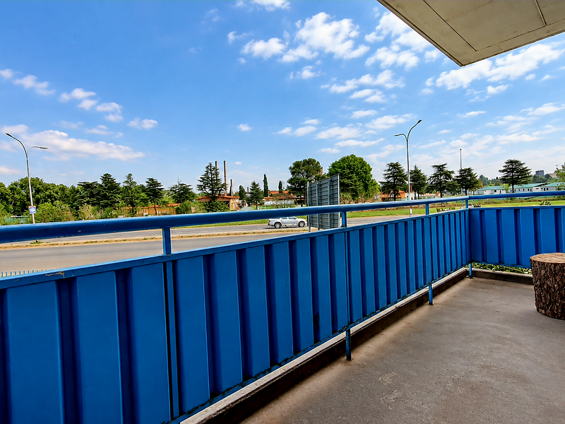 An open balcony featuring a blue metal balustrade and concrete flooring, offering an elevated view of the surrounding street and landscape.