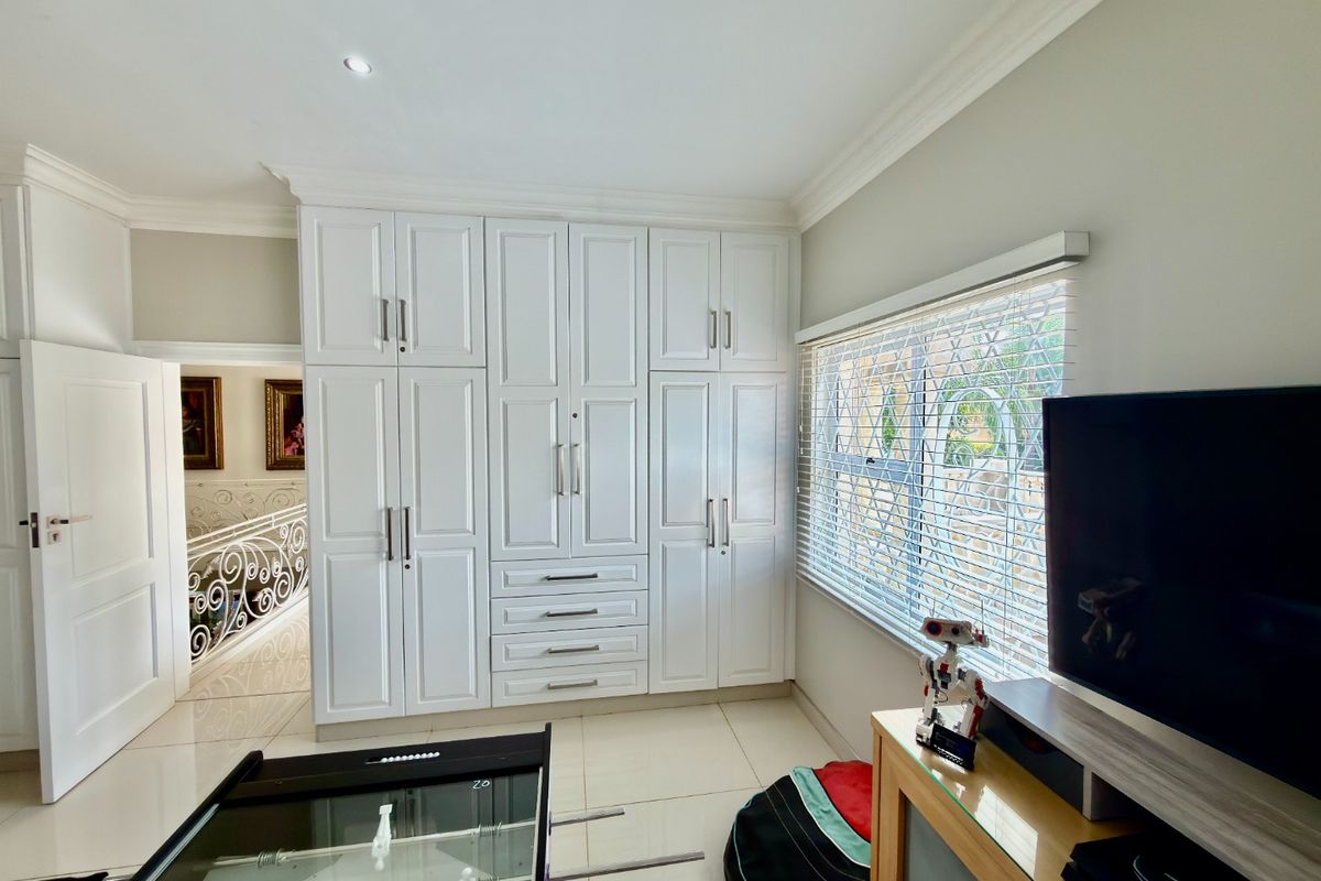 A well-maintained bedroom featuring extensive white built-in cupboards, tiled flooring, and large windows fitted with security bars and venetian blinds.