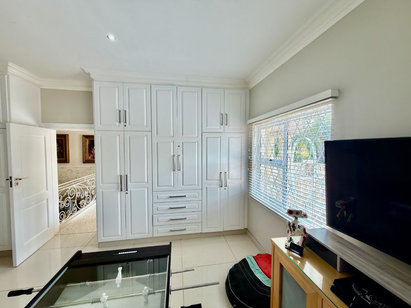 A well-maintained bedroom featuring extensive white built-in cupboards, tiled flooring, and large windows fitted with security bars and venetian blinds.