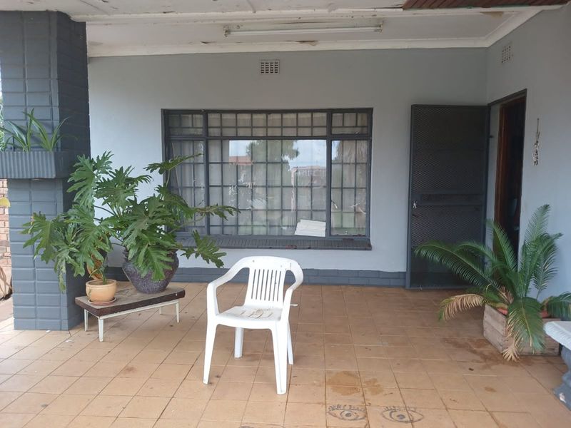 A covered patio area featuring tiled flooring, a security gate, and a large steel-framed window, offering a functional outdoor space in need of cosmetic updates.
