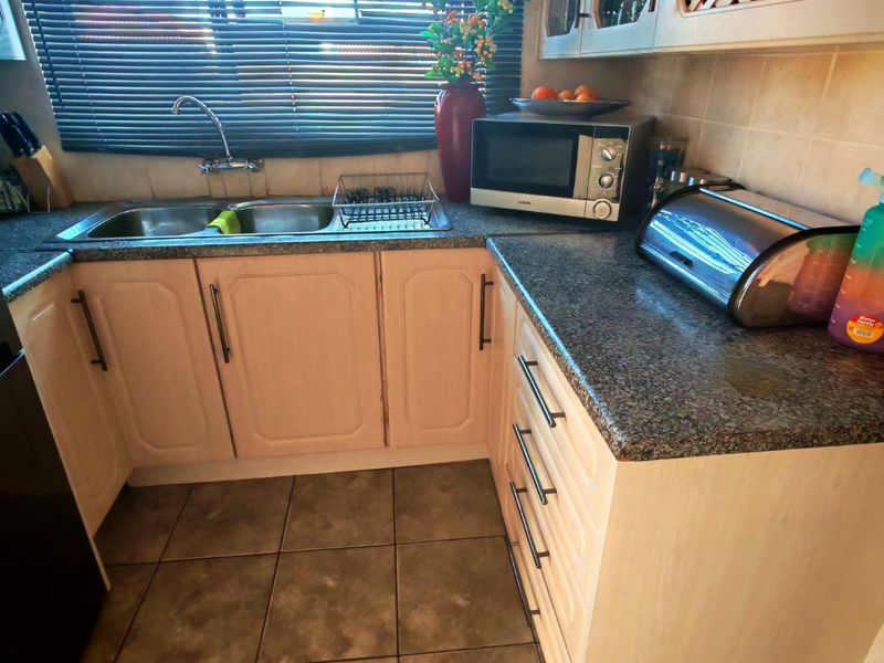 A functional kitchen area featuring laminate cabinetry, granite-look countertops, a double bowl sink, and tiled flooring, illuminated by natural light through venetian blinds.