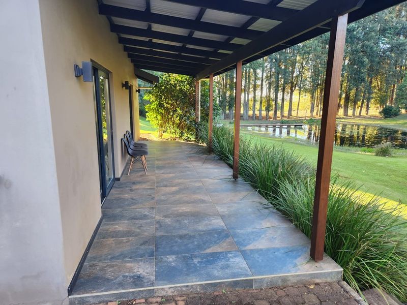 A covered patio featuring large format floor tiles, timber support posts, and a corrugated roof, overlooking a landscaped garden and pond.