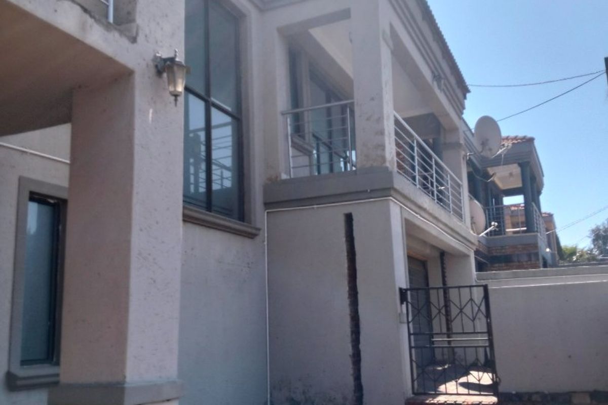 Rear exterior view of a multi-story house featuring a balcony with stainless steel railings, paved ground area, and visible wear on the exterior wall finishes.