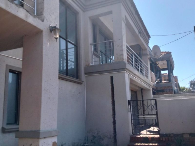 Rear exterior view of a multi-story house featuring a balcony with stainless steel railings, paved ground area, and visible wear on the exterior wall finishes.