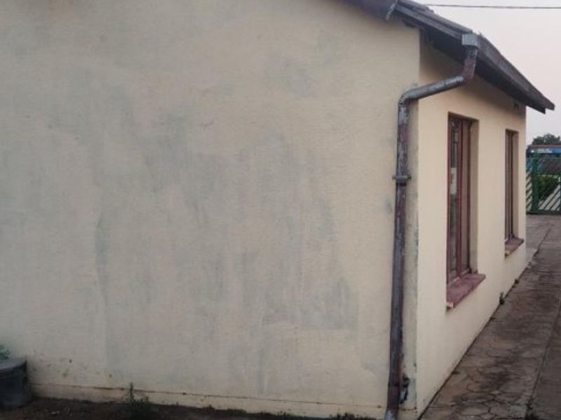 Side view of the property showing exterior wall with visible paint wear, metal downpipe, and concrete apron along the building perimeter.