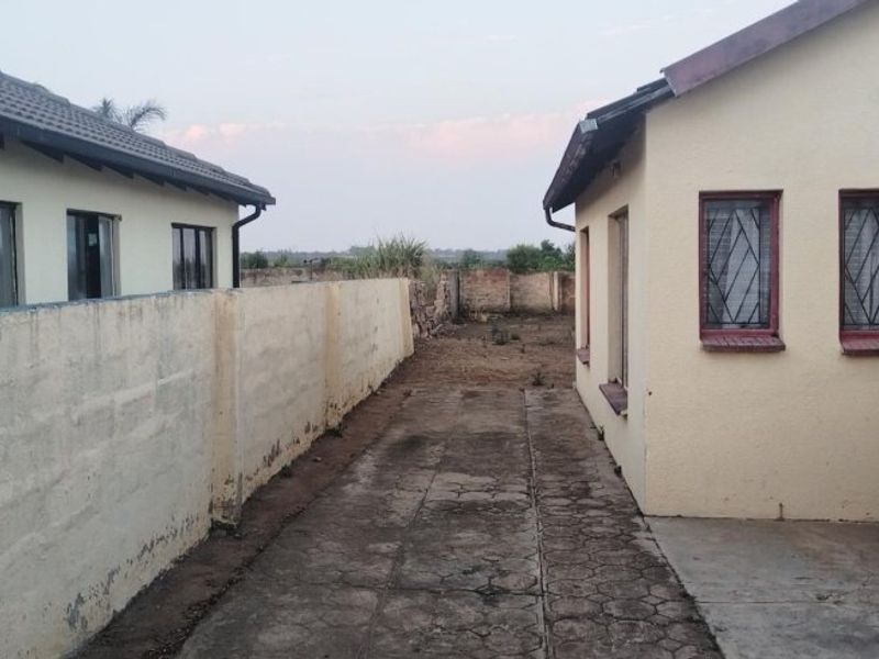 Rear exterior view showing a paved walkway between two residential structures, featuring concrete boundary walls and security-fitted windows.