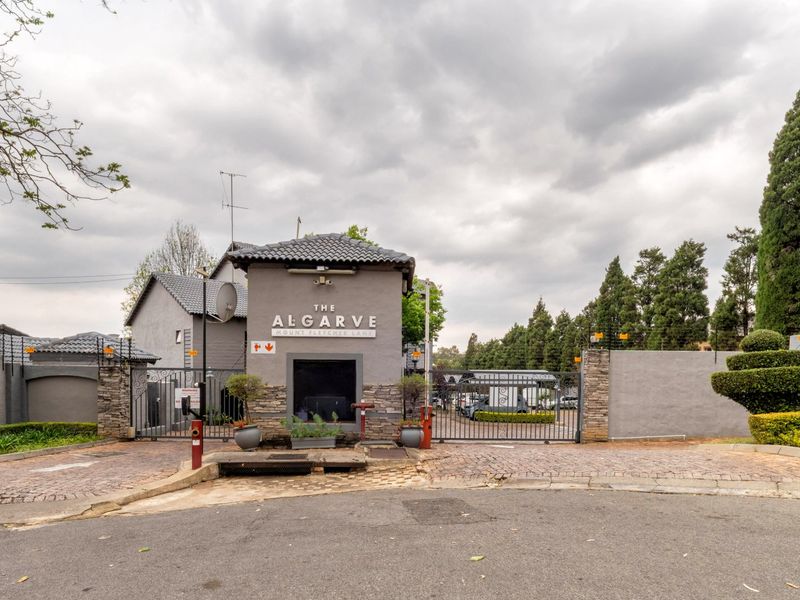 The secure entrance gate and guardhouse of The Algarve complex, featuring stone cladding, tiled roofing, and perimeter security fencing.
