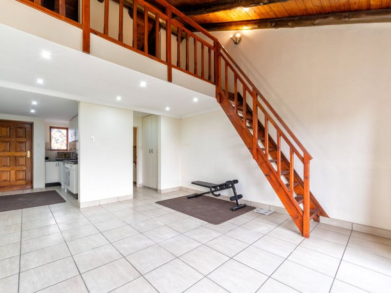 Open-plan living area featuring tiled floors, a wooden staircase leading to a mezzanine level, exposed timber ceiling beams, and recessed lighting.