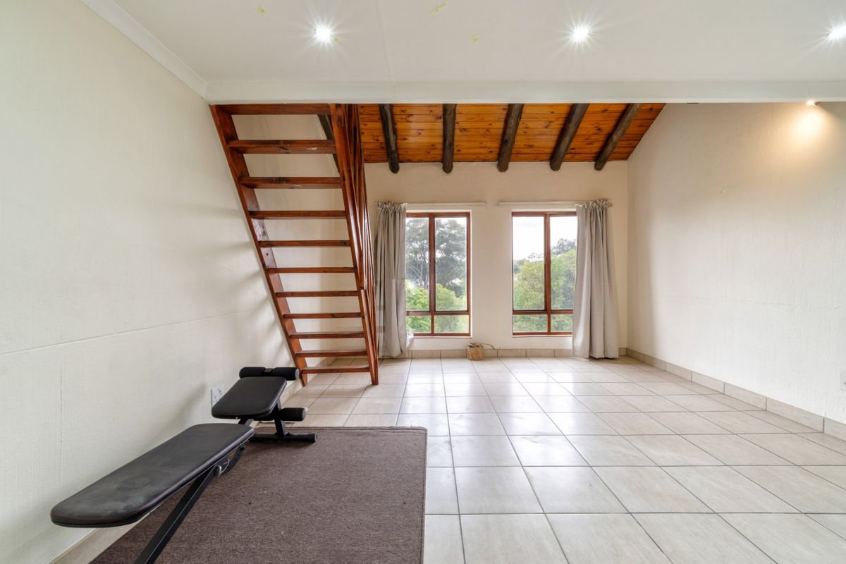 Spacious living area featuring tiled floors, exposed wooden ceiling beams, and a wooden staircase leading to an upper level, with natural light from large windows.