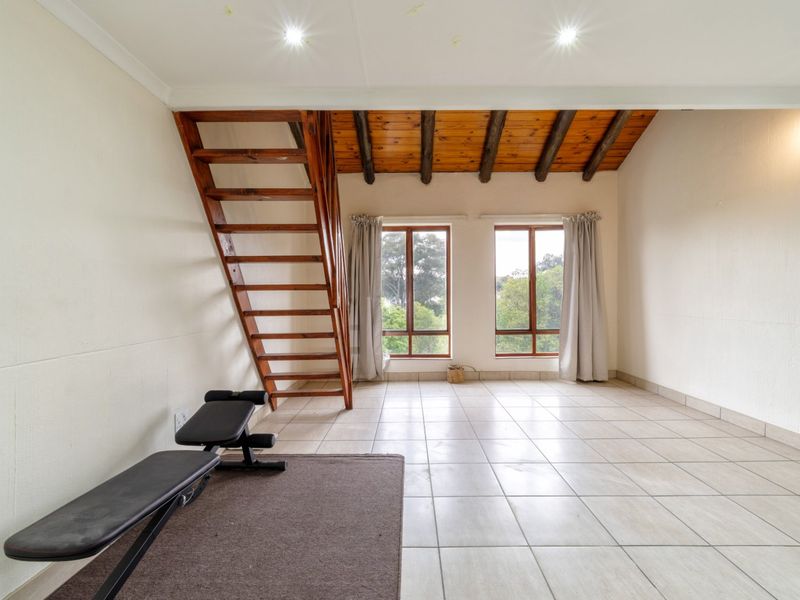 Spacious living area featuring tiled floors, exposed wooden ceiling beams, and a wooden staircase leading to an upper level, with natural light from large windows.