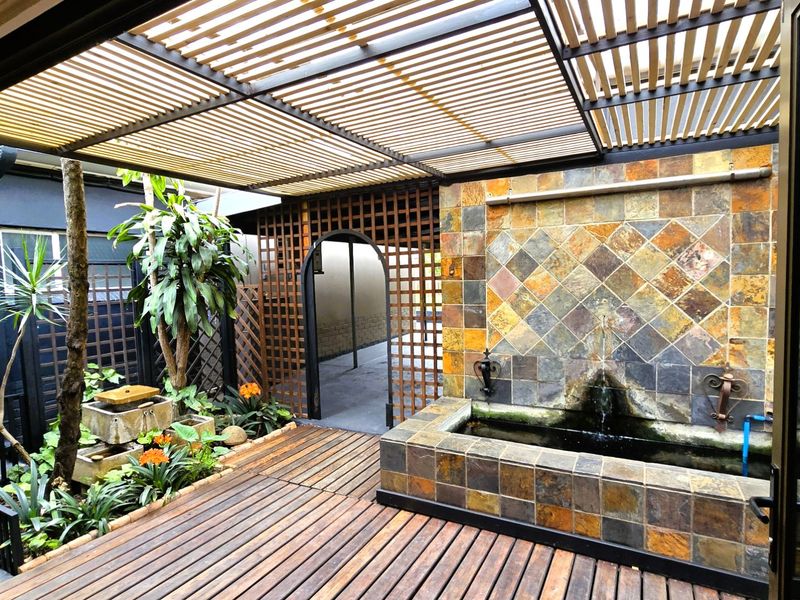 A covered outdoor patio featuring timber decking, a slatted pergola, and a decorative stone-tiled water feature, creating a tranquil courtyard atmosphere.