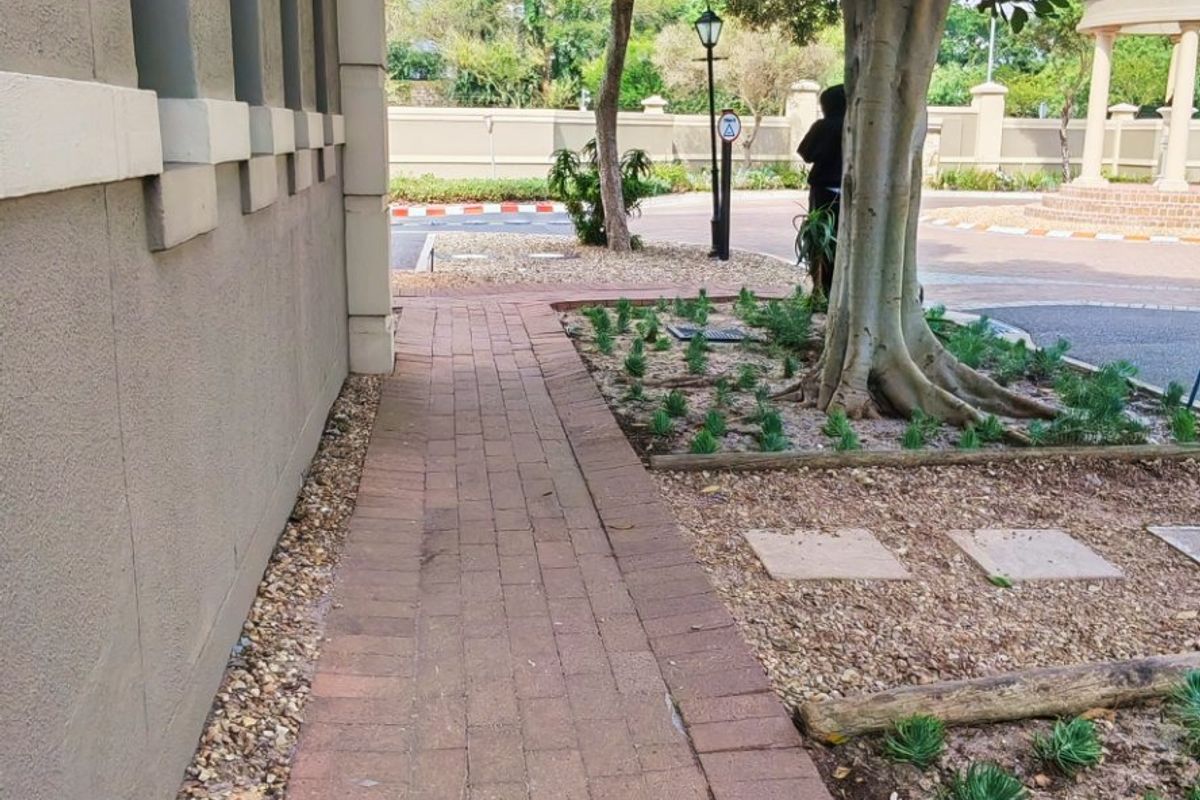A paved brick walkway runs alongside the exterior wall of the apartment building, bordered by landscaped garden beds and mature trees.