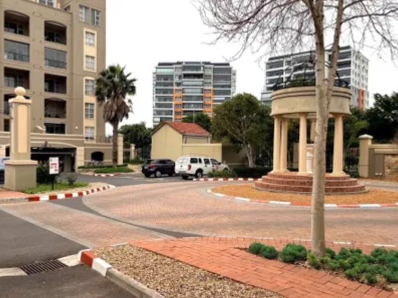 A view of the apartment complex entrance featuring a paved circular driveway, landscaped garden beds, and a decorative central rotunda structure.