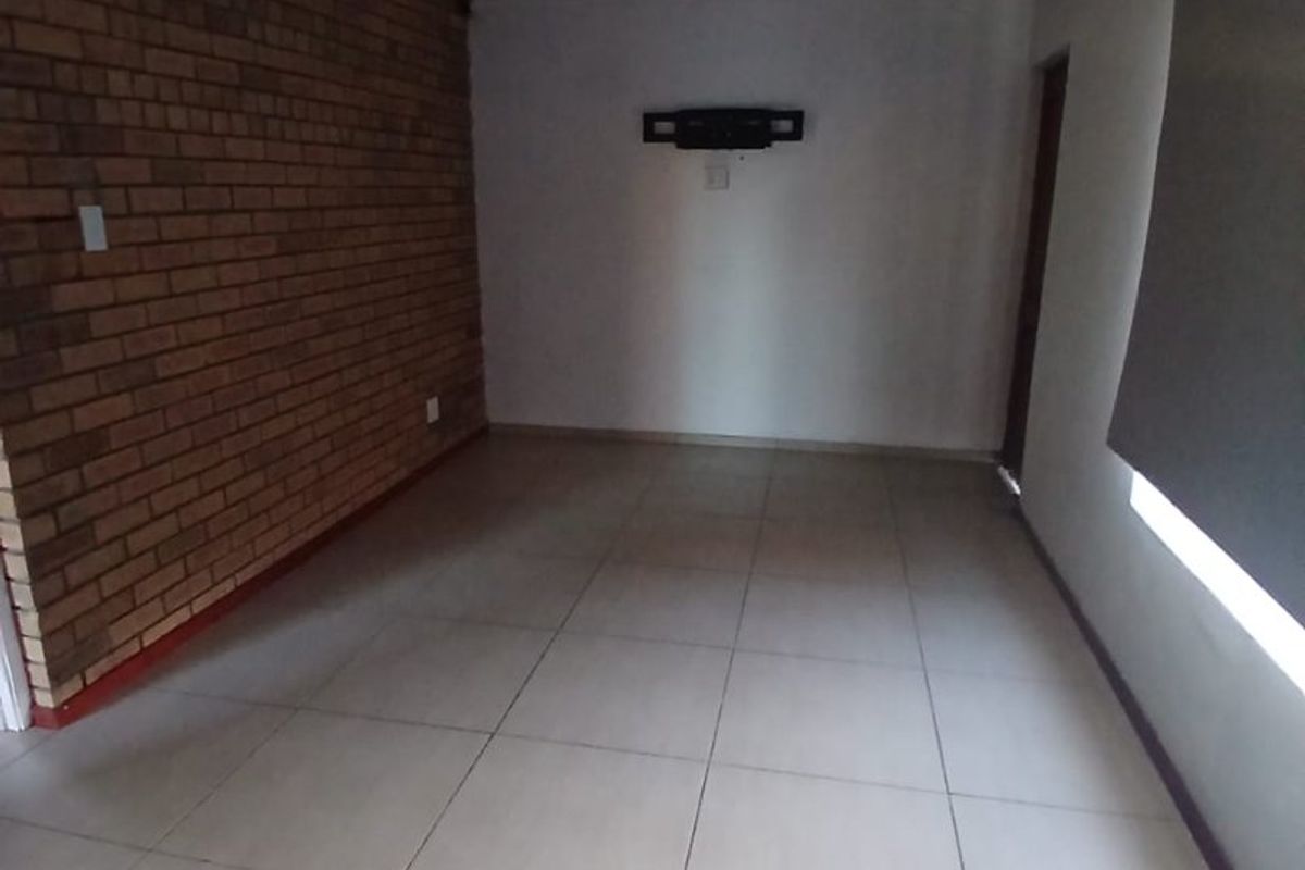 An empty living room featuring an exposed brick accent wall, neutral ceramic floor tiles, and a roller blind.