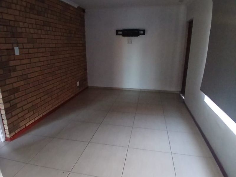 An empty living room featuring an exposed brick accent wall, neutral ceramic floor tiles, and a roller blind.