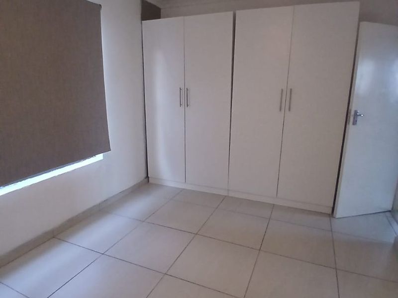 A clean, empty bedroom featuring neutral tiled flooring, built-in white cupboards, a ceiling fan, and a roller blind.