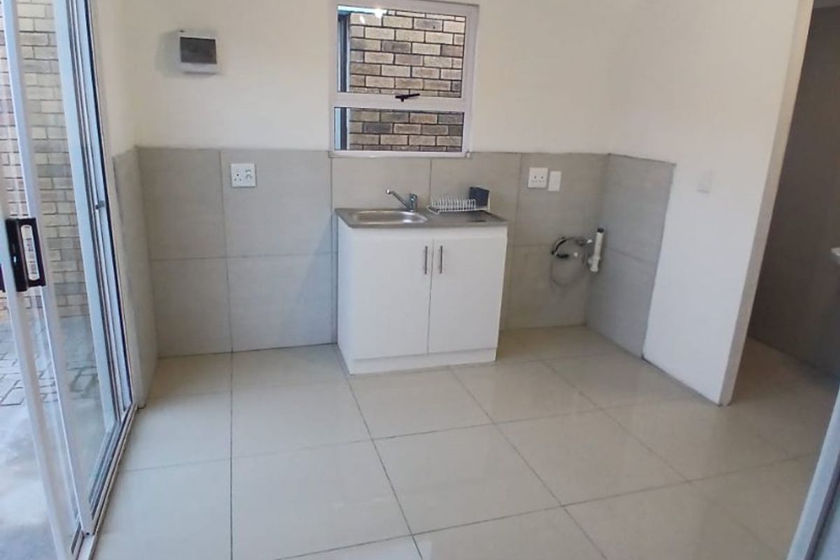 A clean, empty kitchen area featuring tiled flooring, a built-in sink with splashback, and a sliding glass door providing access to the exterior.