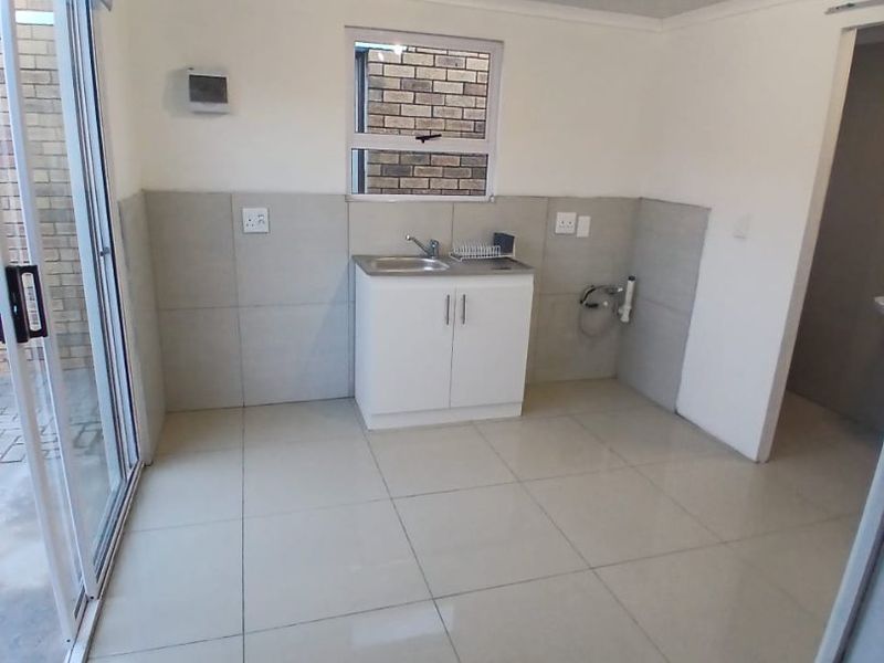 A clean, empty kitchen area featuring tiled flooring, a built-in sink with splashback, and a sliding glass door providing access to the exterior.