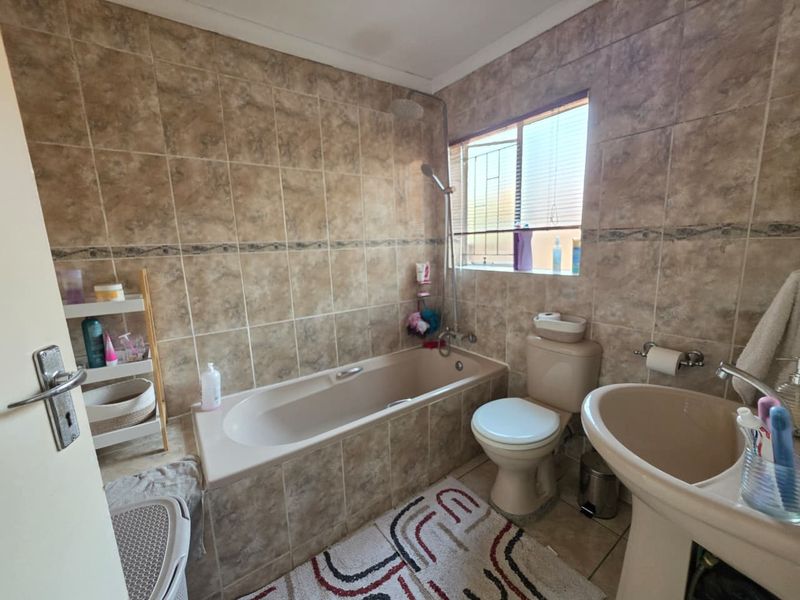 A functional bathroom featuring a built-in bathtub with shower attachment, pedestal basin, and toilet, finished with neutral-toned wall and floor tiles.