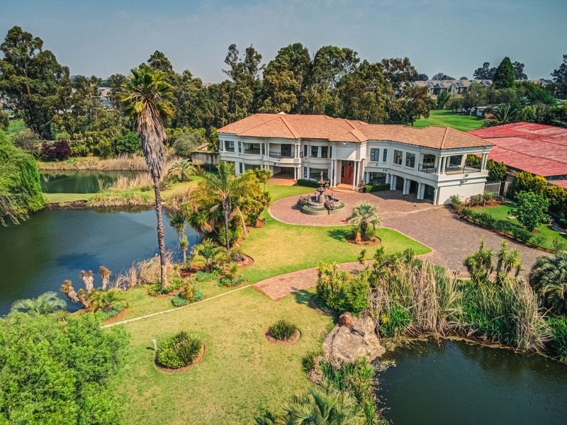 Aerial view of a large luxury residence featuring a paved driveway, central water fountain, landscaped gardens, and a scenic pond setting.