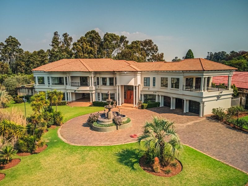 An aerial view of a grand double-story residence featuring a paved circular driveway, central stone fountain, manicured gardens, and classic architectural detailing.
