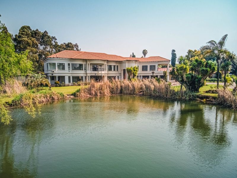 A spacious multi-level residence featuring a terracotta tile roof, expansive windows, and a balcony, set against a scenic pond and landscaped garden.