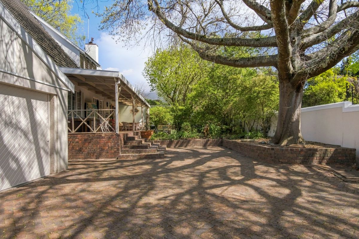 A spacious rear garden featuring a brick-paved patio, a covered veranda, and a mature tree set within a landscaped area with brick retaining walls.