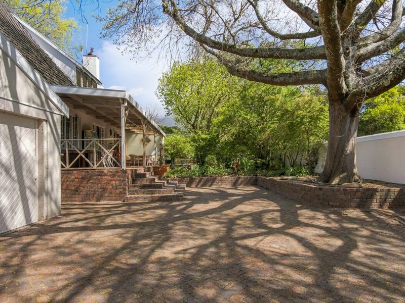 A spacious rear garden featuring a brick-paved patio, a covered veranda, and a mature tree set within a landscaped area with brick retaining walls.