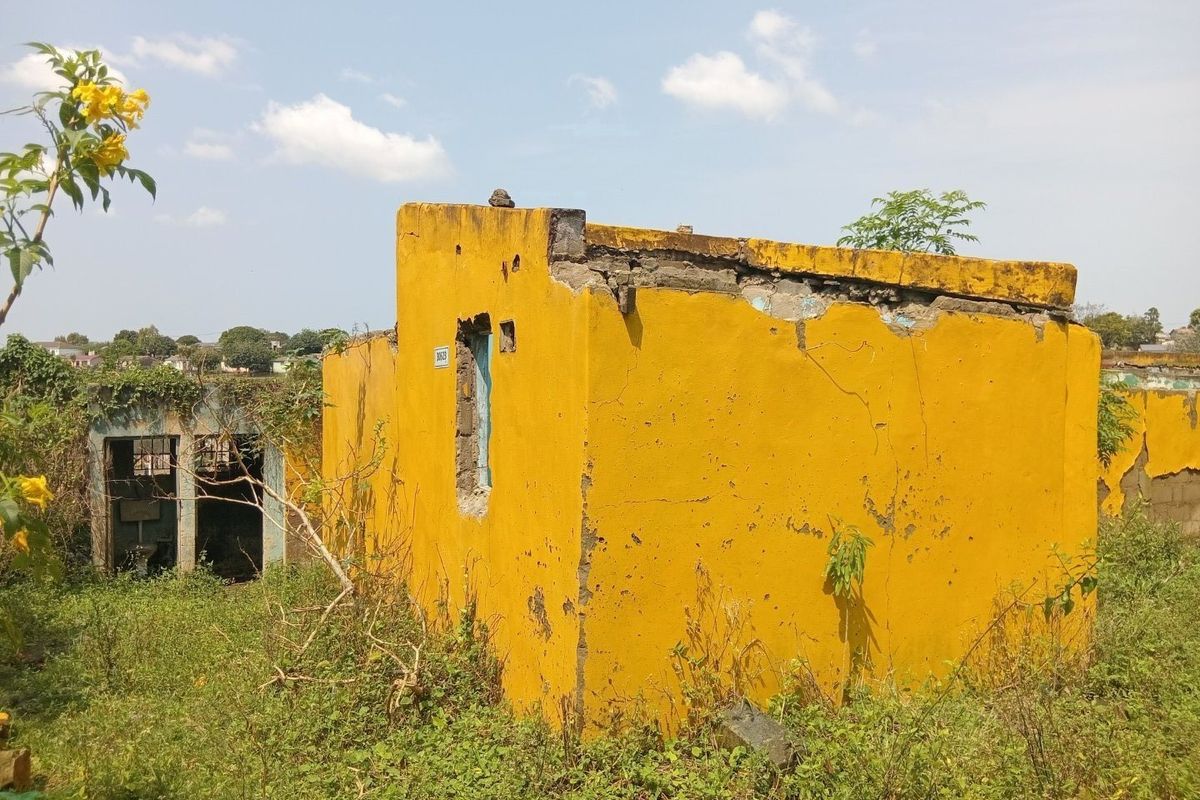 A dilapidated exterior structure showing significant masonry damage, crumbling walls, and severe structural deterioration in an overgrown outdoor setting.