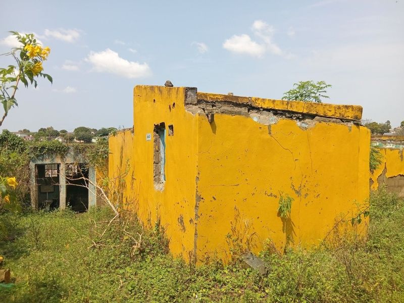 A dilapidated exterior structure showing significant masonry damage, crumbling walls, and severe structural deterioration in an overgrown outdoor setting.