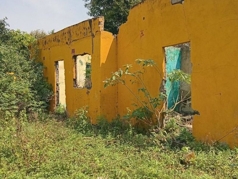 A severely dilapidated structure showing significant structural failure, missing roof, and extensive wall damage, surrounded by overgrown vegetation.