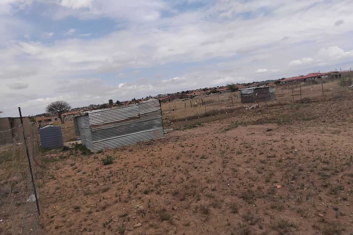 A vacant residential plot featuring wire fencing, a water storage tank, and basic corrugated metal structures in a suburban setting.