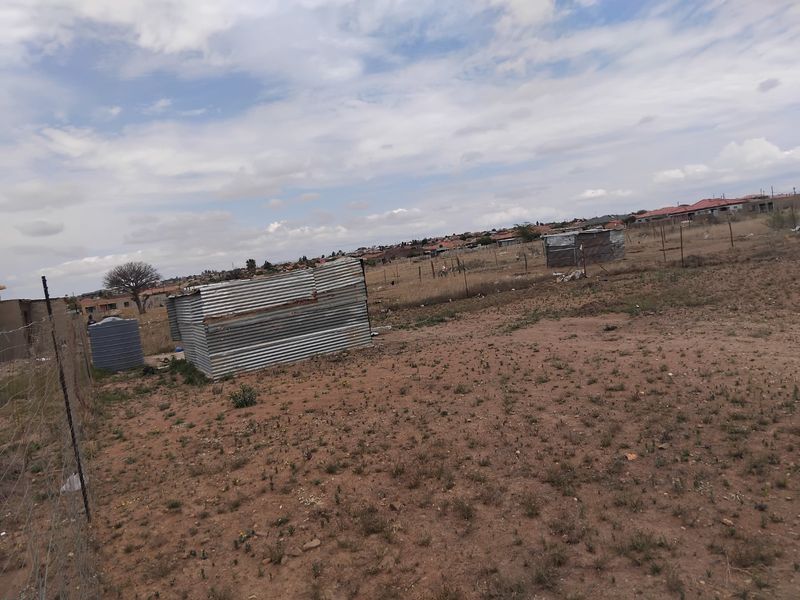 A vacant residential plot featuring wire fencing, a water storage tank, and basic corrugated metal structures in a suburban setting.