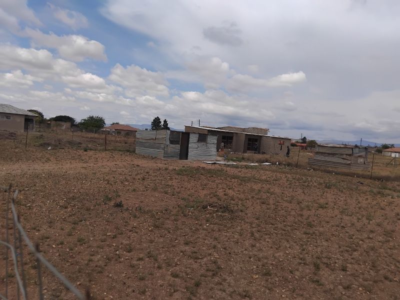 A vacant residential plot featuring unpaved ground, wire fencing, and several small, unfinished corrugated metal structures.