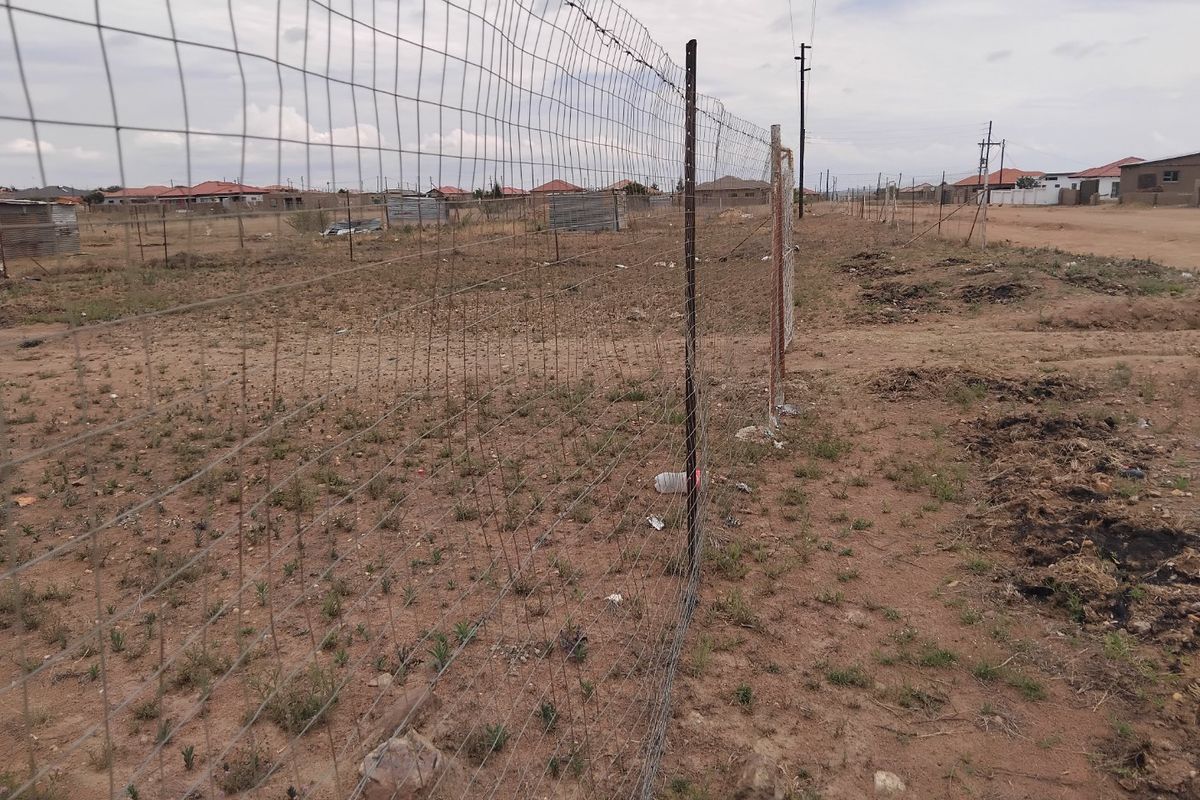 A view of a vacant residential plot featuring wire mesh perimeter fencing and unlandscaped ground in a developing neighborhood.