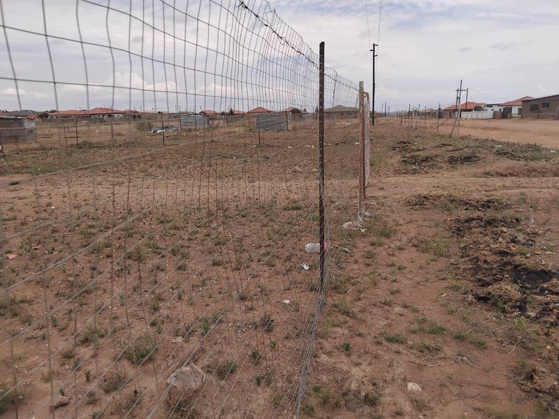 A view of a vacant residential plot featuring wire mesh perimeter fencing and unlandscaped ground in a developing neighborhood.