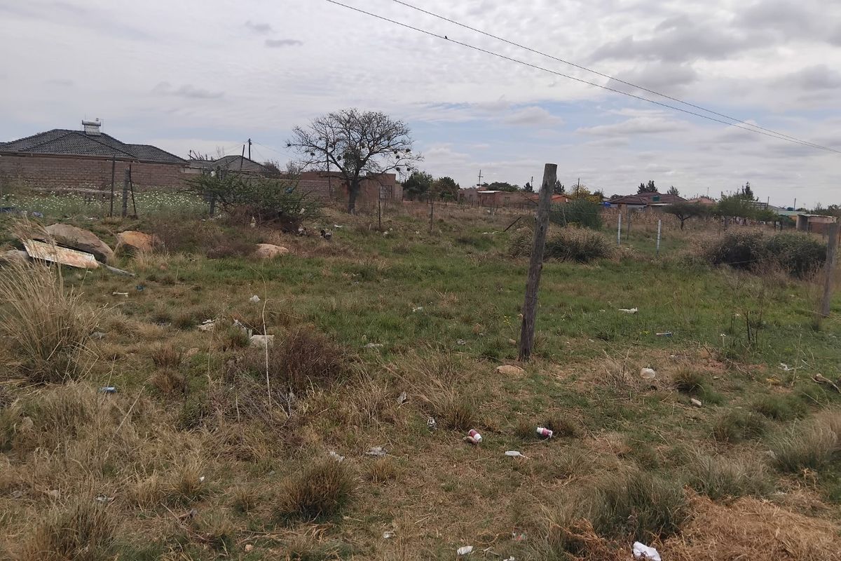 A vacant residential plot featuring natural grass, scattered debris, and basic wire fencing with overhead power lines visible.